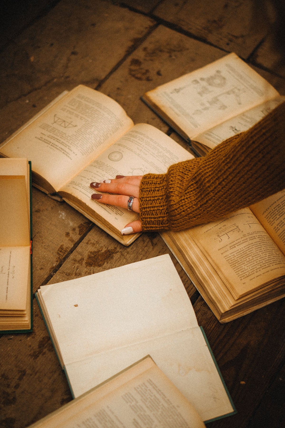 A hand rests on an open book among several aged volumes on a wooden table.